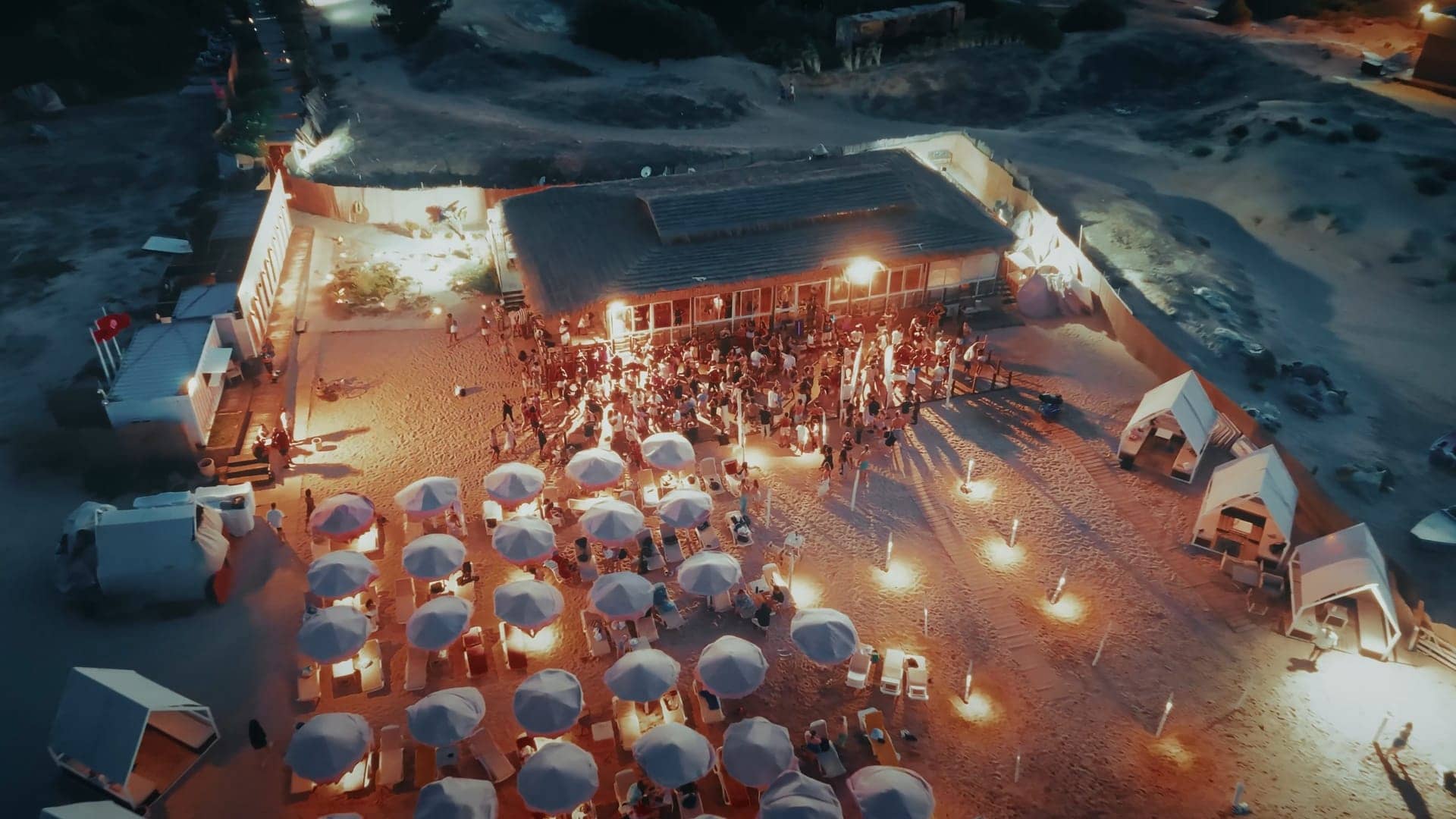Aerial view of the beachfront party at dusk, umbrellas and festival lights below